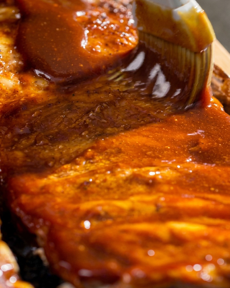 刷毛を使って、焼き上がったスペアリブの表面にツヤのある赤いBBQソースをたっぷりと塗っている場面。