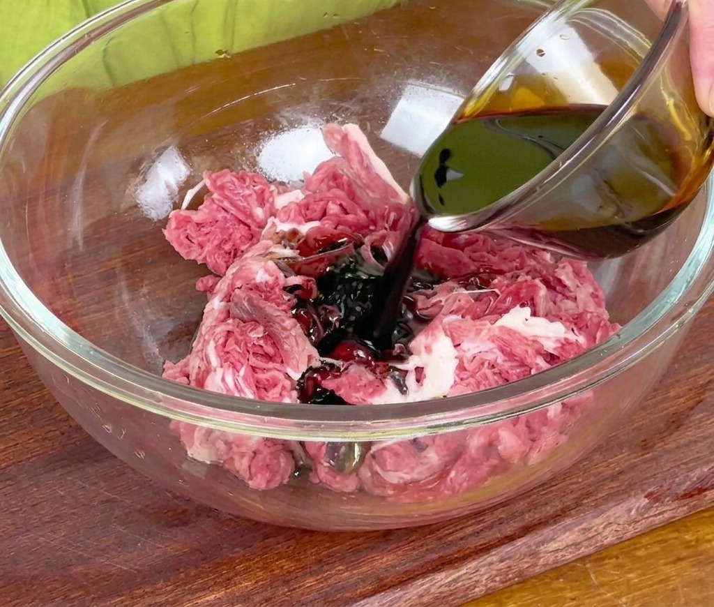 大きなガラスボウルに入った牛肉に、小さなガラスの器から醤油を注いでいる様子。