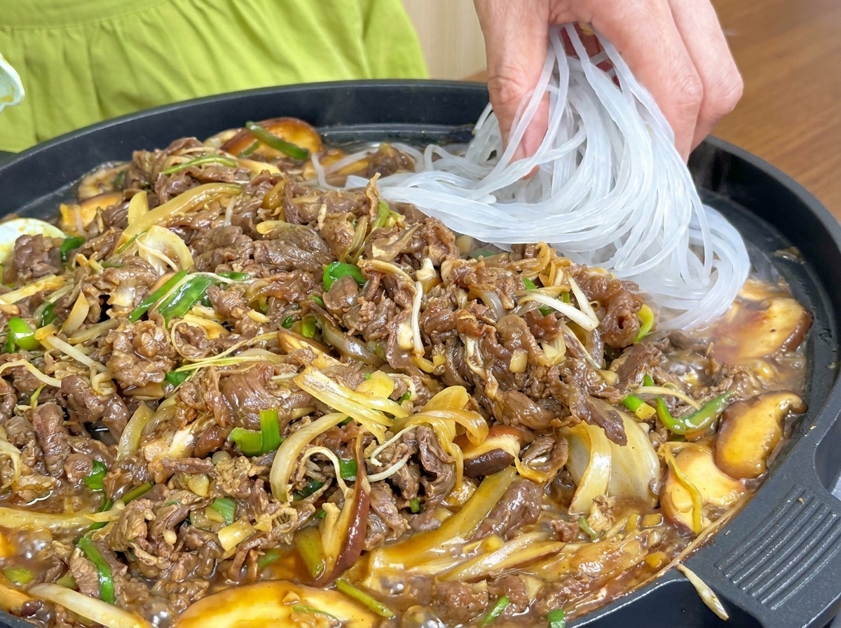 調理中の牛肉と野菜の入ったフライパンに、半透明の春雨を加えている様子。