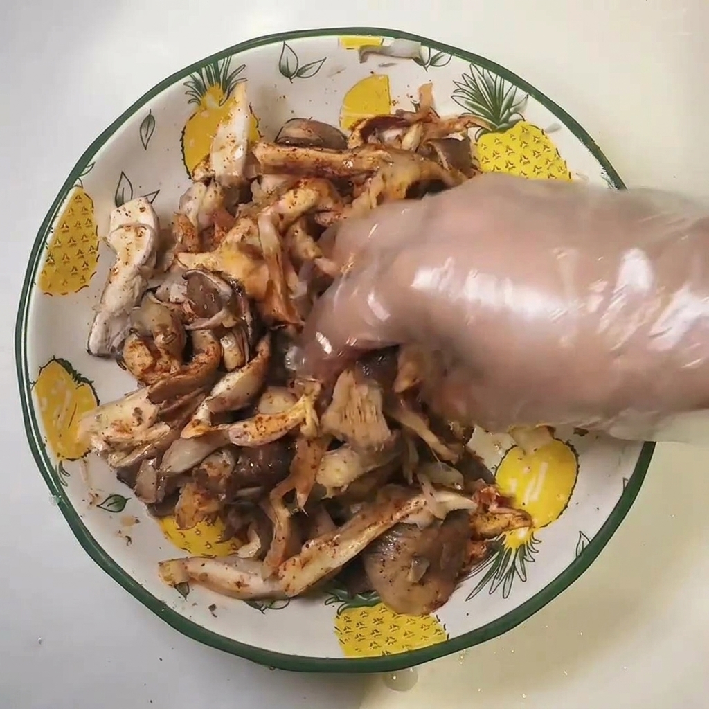 手袋をした手で、ボウルに入った味付け済みのひらたけを混ぜ合わせている様子。