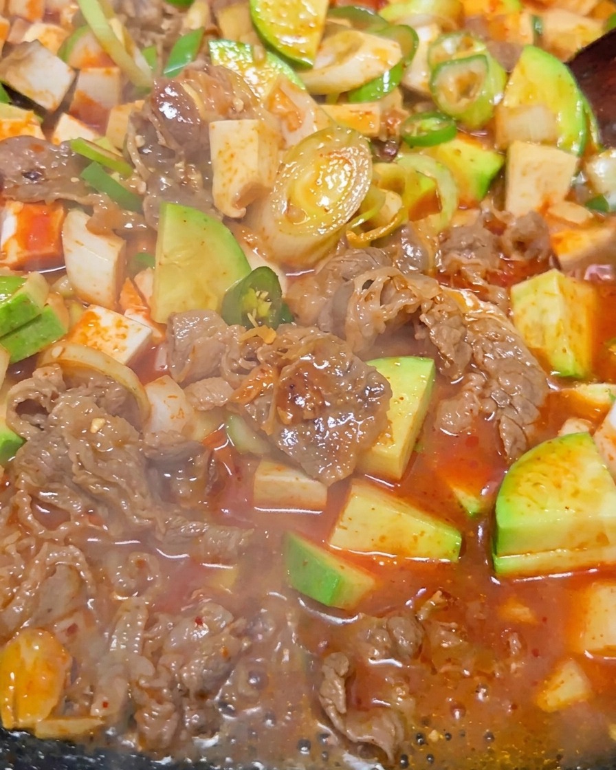 赤茶色のスープの中で豆腐、牛肉、緑のズッキーニがグツグツと激しく煮立っている、韓国風味噌チゲのクローズアップ。