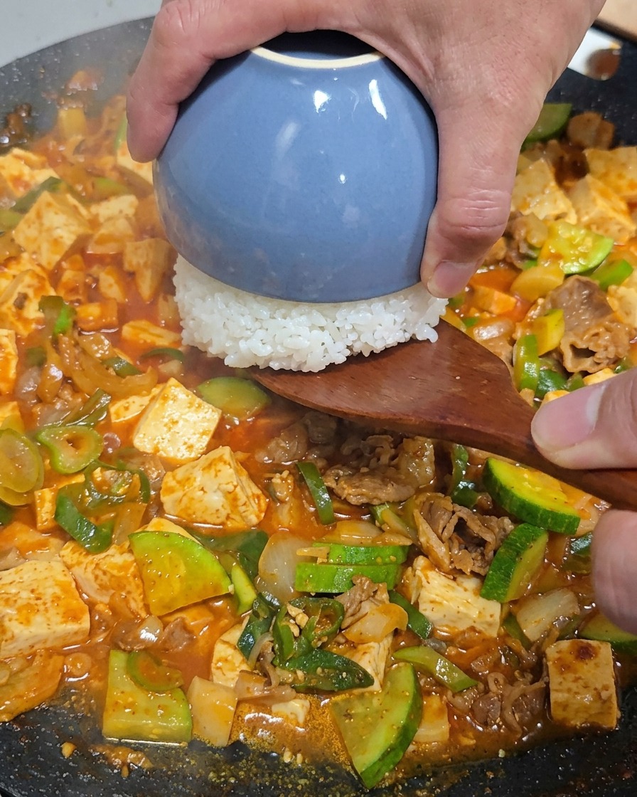 牛肉と野菜のチゲが入った鍋の中央に、木べらを使って青い小鉢に入った白米をひっくり返して盛り付けている様子。