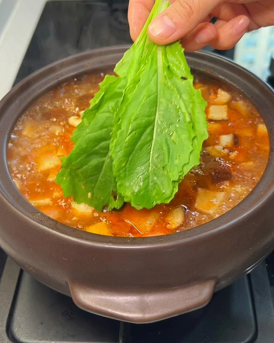 手を使って、新鮮な緑の葉物野菜を煮立っているチゲに加えている様子。