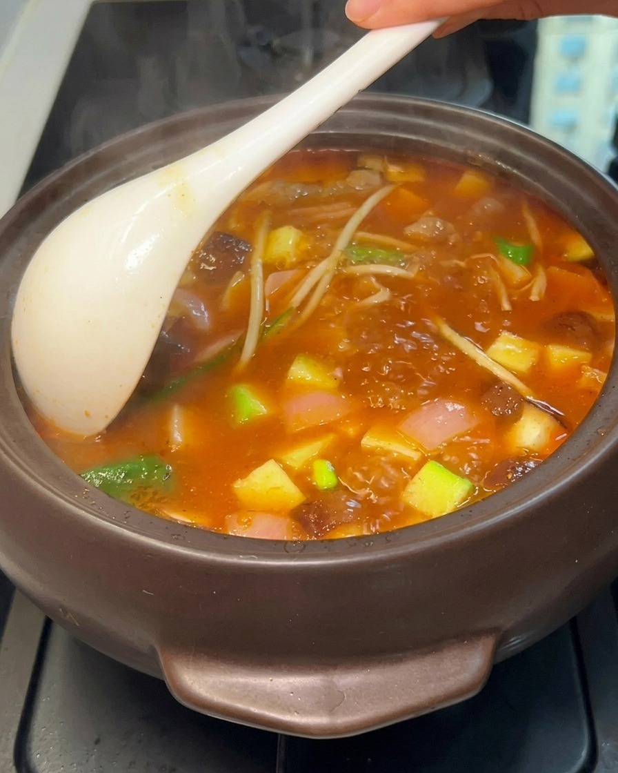 煮立った鍋を白いお玉で優しくかき混ぜ、豆腐、ズッキーニ、キノコ、牛肉が赤いスープに混ざり合っている様子。