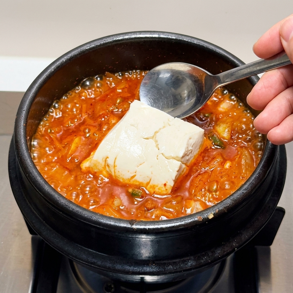 沸騰するキムチチゲの中に、大きなスプーンで絹ごし豆腐を崩さないように入れている様子。