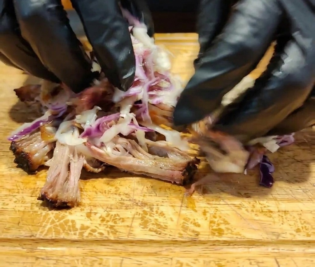 手袋をした手で、裂いた肉、濃厚なソース、色鮮やかなコールスローをカッティングボードの上で混ぜ合わせている様子。