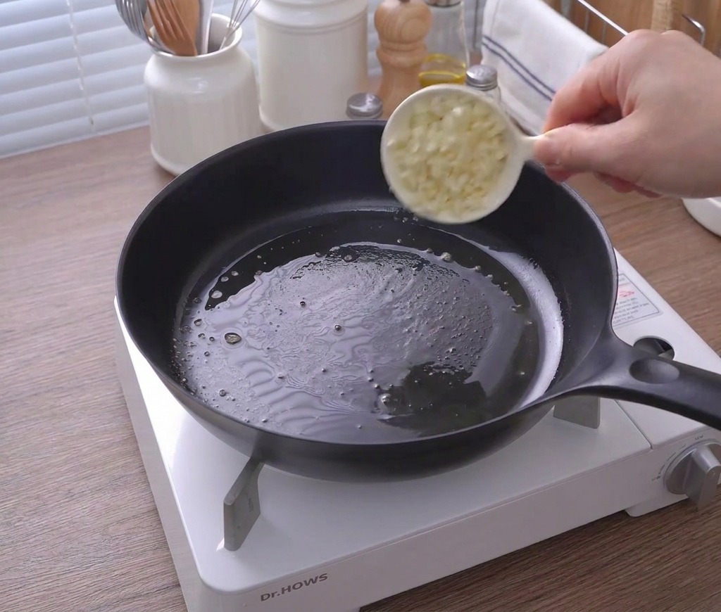 溶けたバターが入ったフライパンに、白いカップからみじん切りのにんにくを入れている様子。