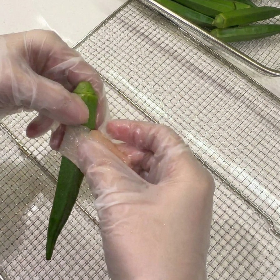 手袋をはめた手で、薄切りにした鶏肉をオクラに螺旋状に巻きつけている様子。