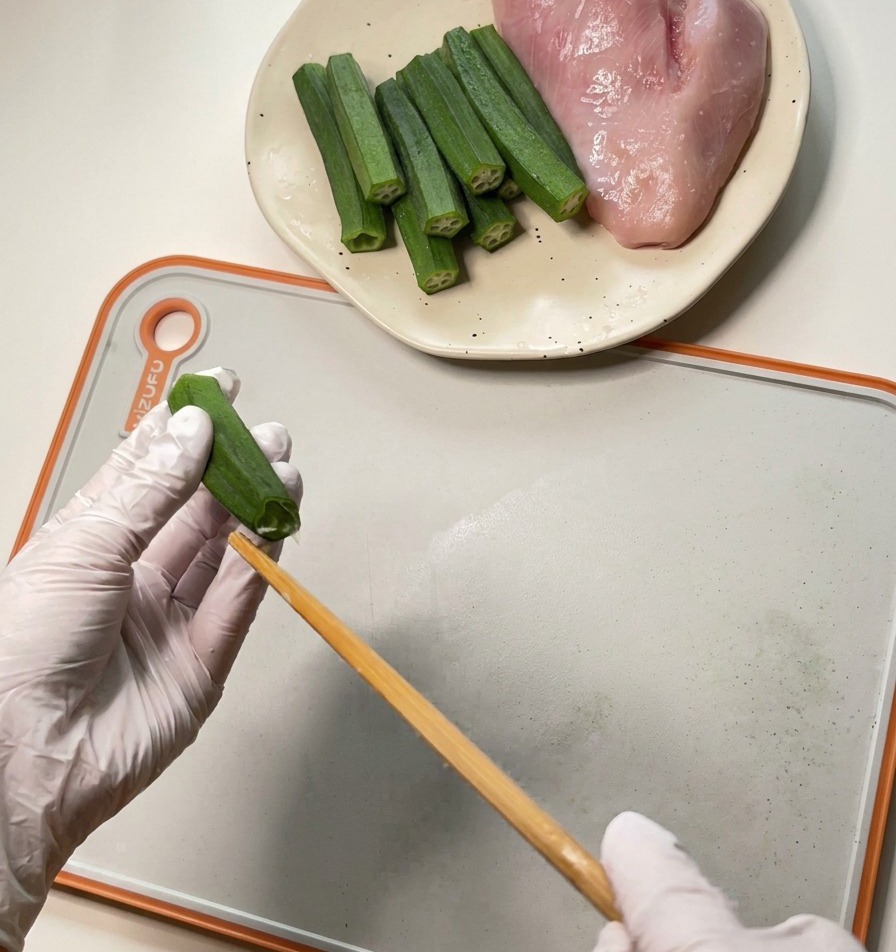 白い手袋をはめた人が、木製の箸を使ってオクラの中から種とワタを押し出している様子。
