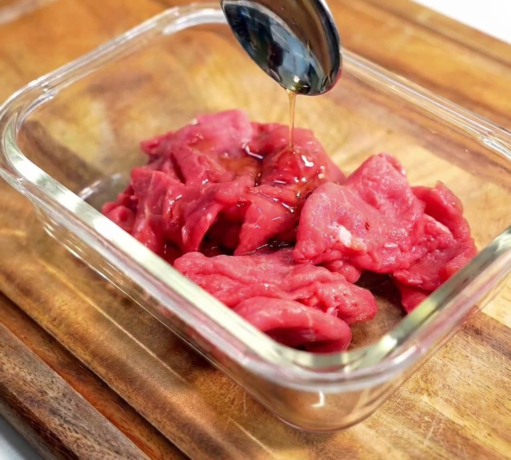 木製の台の上で、ガラスボウルに入ったスライス肉に調味料を回しかけている様子