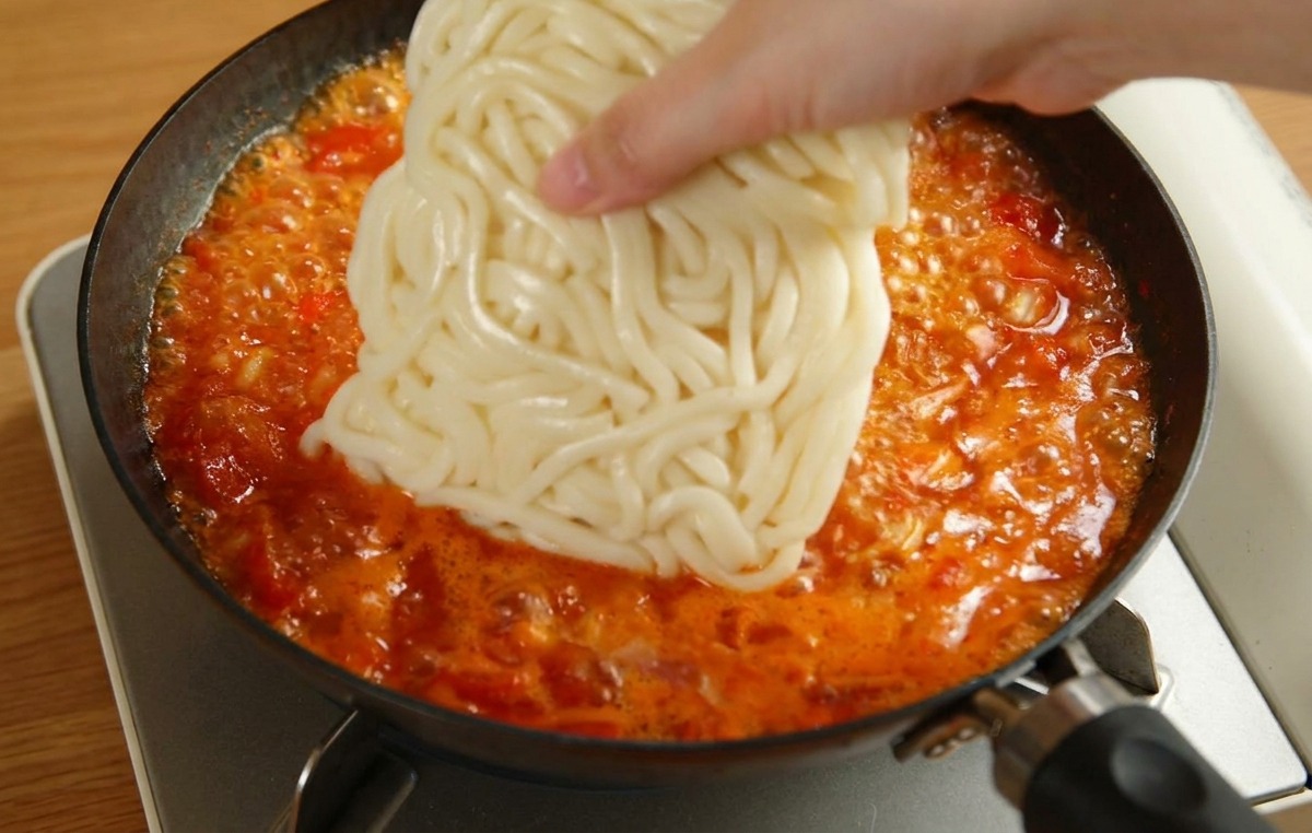 煮え立つ赤いトマトソースの中に、白い冷凍うどんの塊を入れている手元の様子。