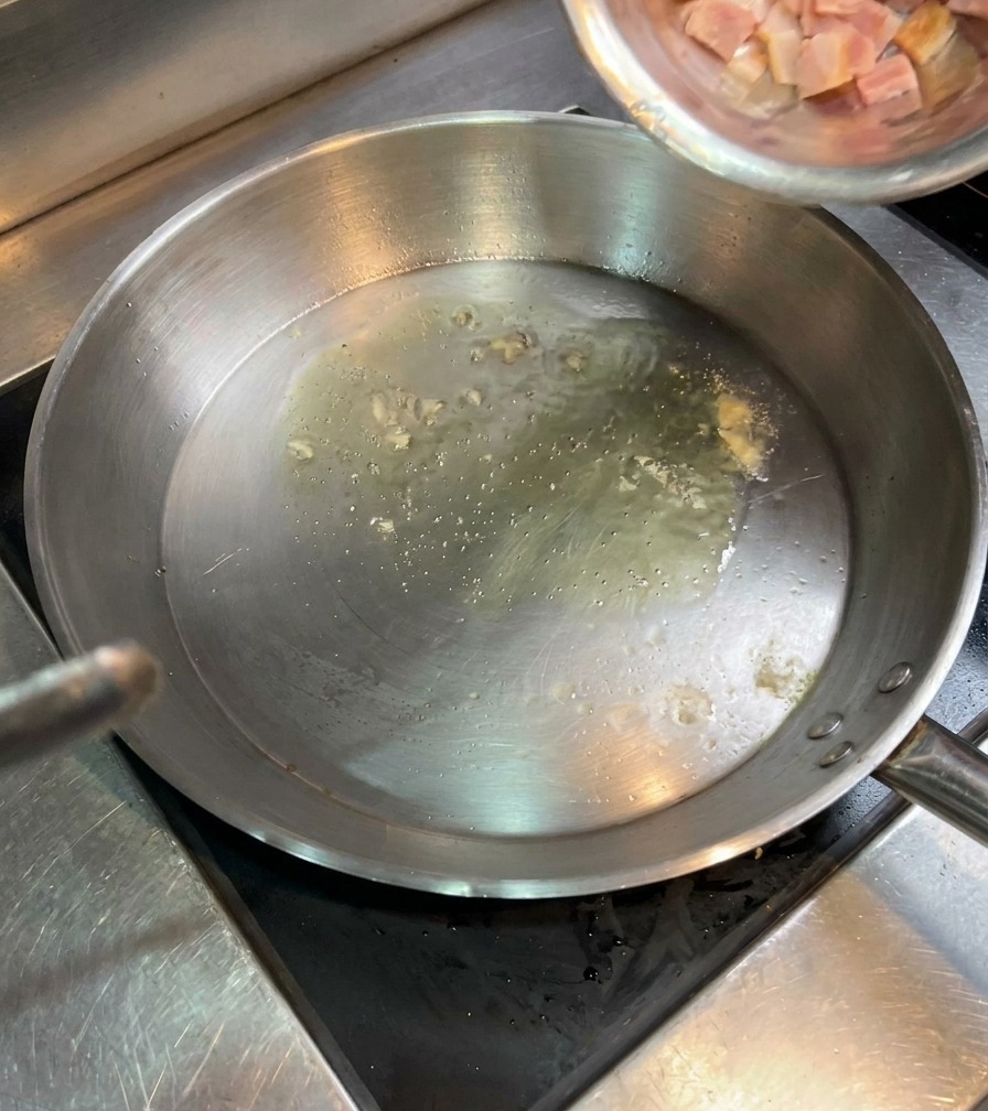 熱い油が入ったフライパンに、ステンレスボウルから角切りベーコンを入れている様子。