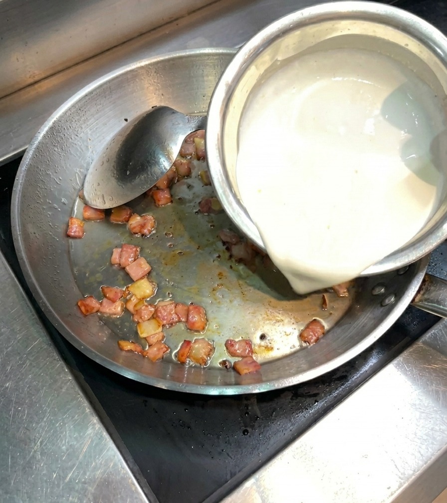 カリカリに炒めたベーコンが入ったフライパンに、白い生クリームを注いでいる様子。