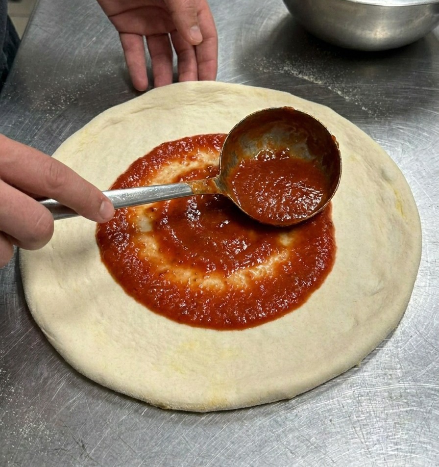 平らに伸ばしたピザ生地の中心に、レードルを使って鮮やかな赤色のトマトソースを円を描くように広げている様子。