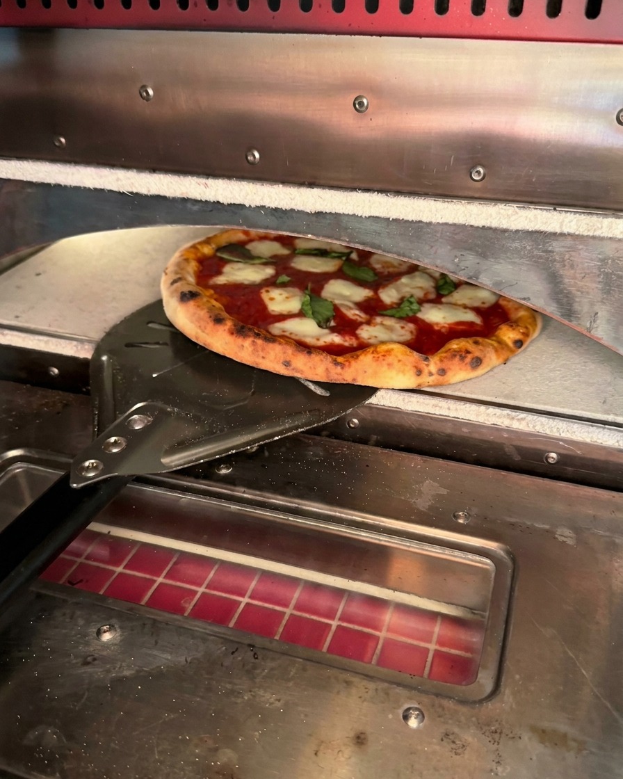 縁にこんがりと焦げ目がついた焼き立てのピザを、金属製のピールで窯から取り出している様子。