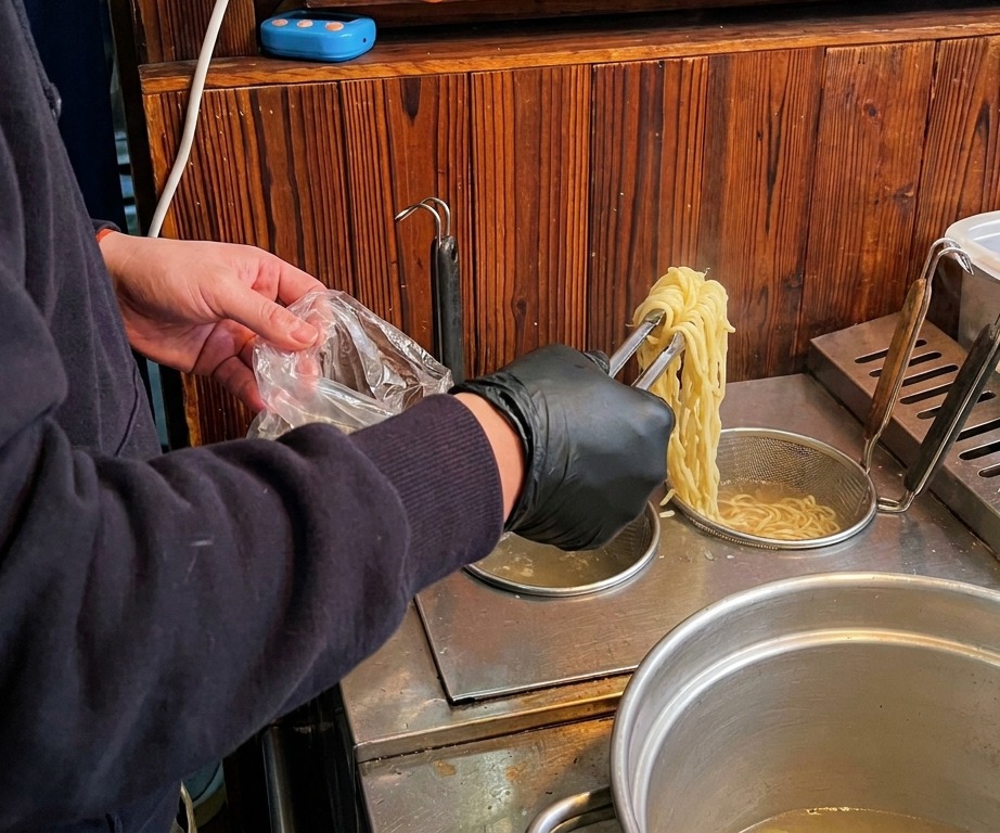 沸騰した鍋からトングで黄色い中華麺を引き上げ、丼に移す準備をしている様子。