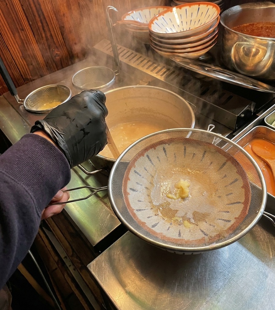 湯気が立つクリーミーな白いスープを、こし器を通してラーメン鉢に注いでいる様子。