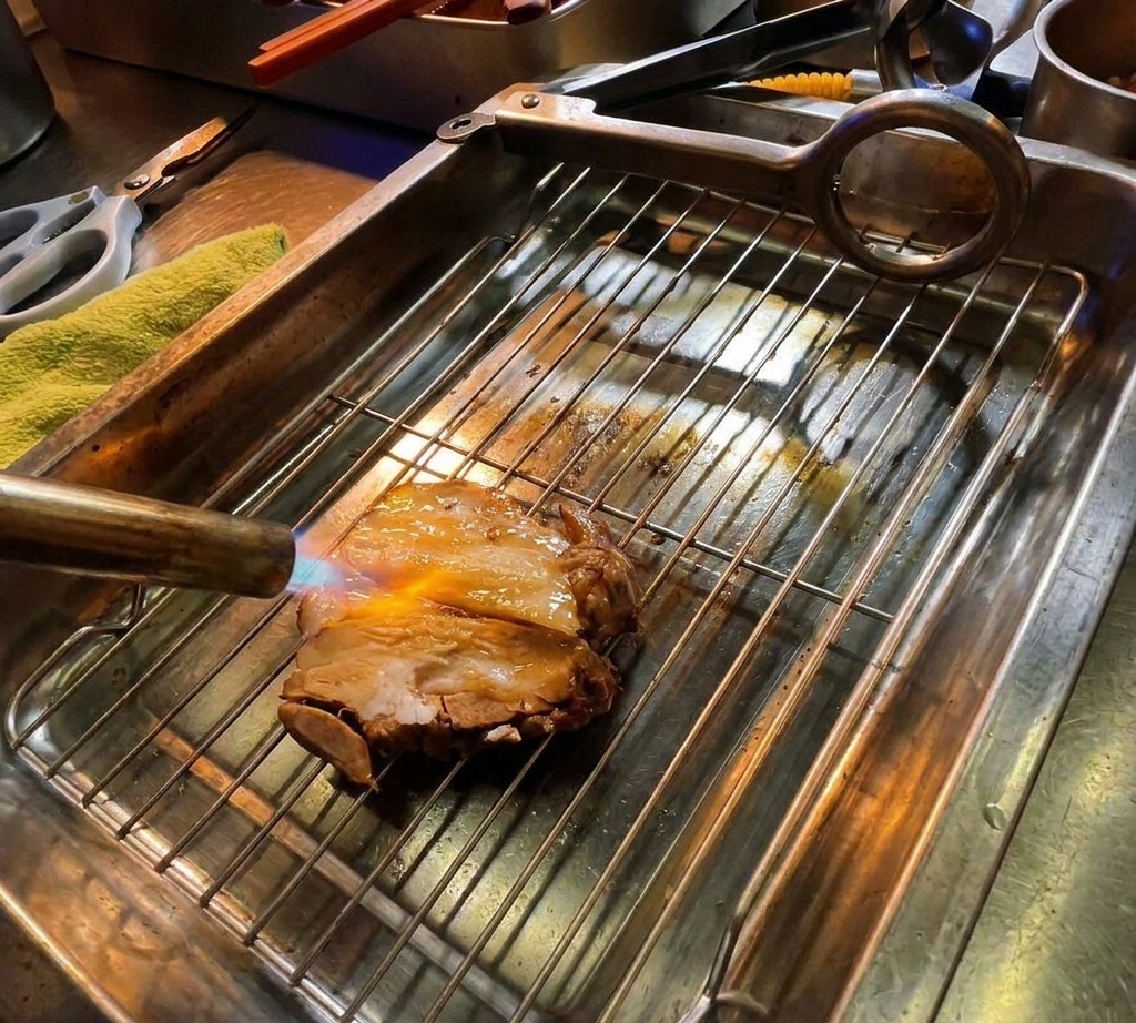 キッチンバーナーの炎で、網の上の厚切りチャーシューに焼き色をつけている様子。