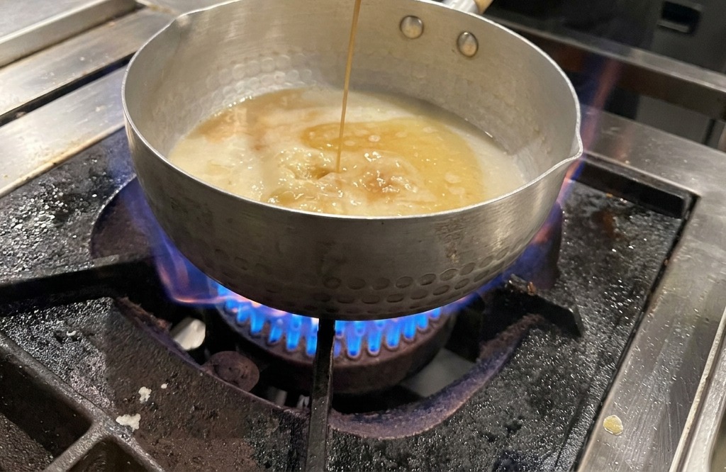 ガスコンロの火にかかったスープに、レードルから濃い色の醤油が注がれている様子。
