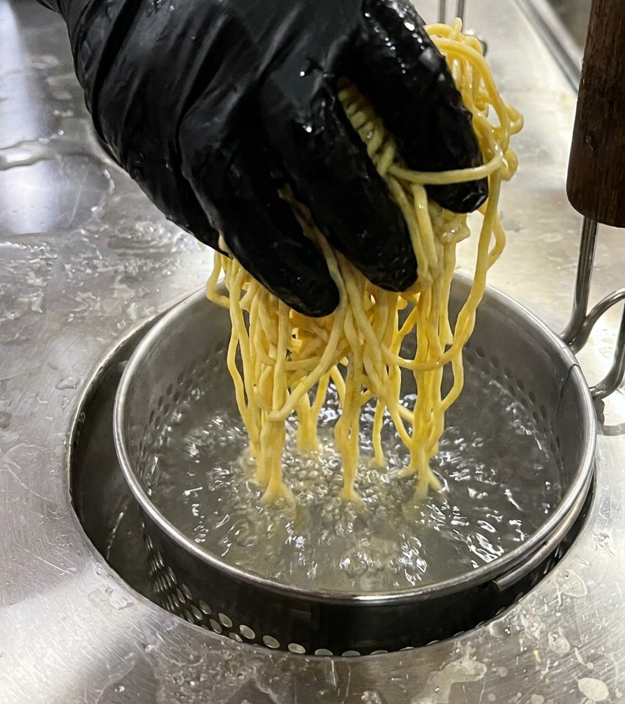 テボ（振りザル）に入った黄色の生麺を、たっぷりとした沸騰したお湯に入れる様子。