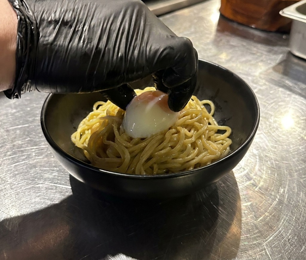 茹で上がった麺の上に、手袋をした手で慎重にプルプルの温泉卵をのせている様子。