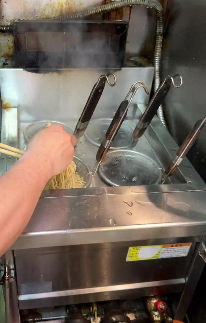 沸騰した鍋の中で、テボ（麺揚げザル）に入った麺を箸でほぐしながら茹でている様子。