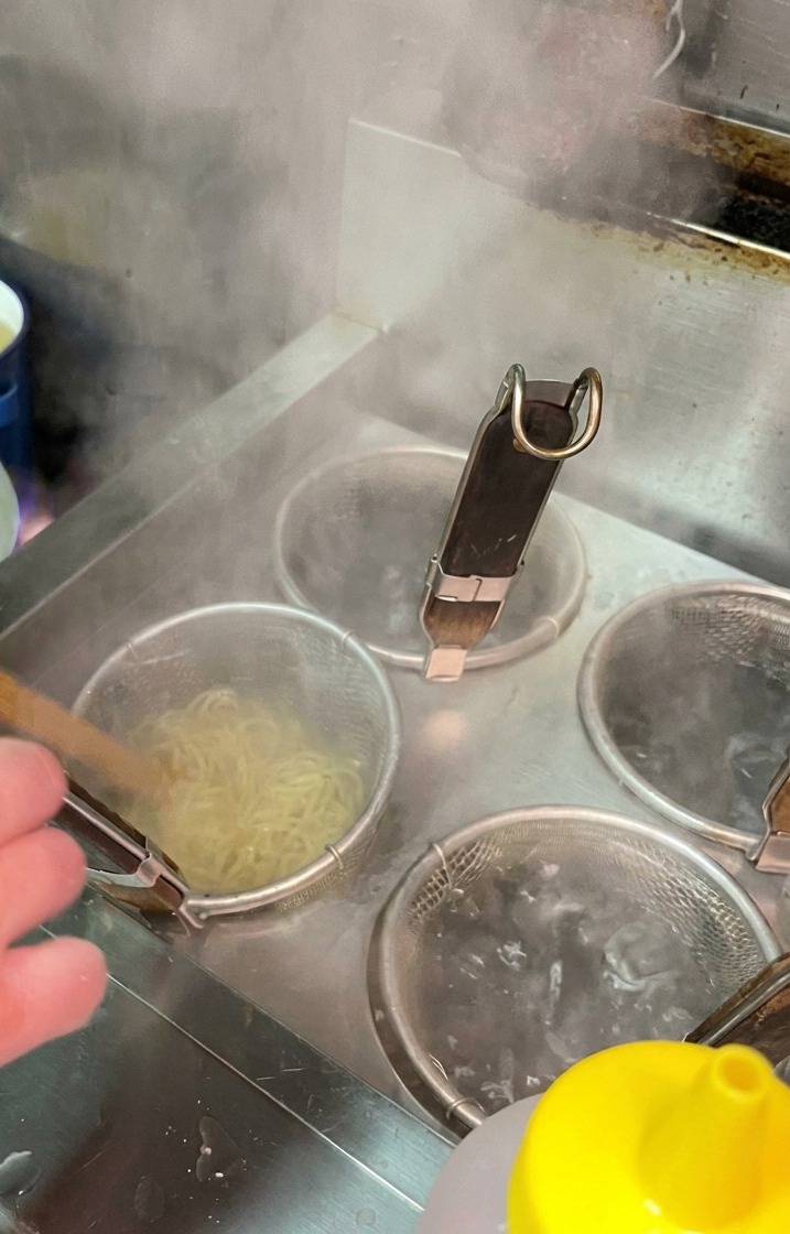 プロの厨房で、茹で上がった麺が入ったテボを勢いよく振って湯切りをしている様子。