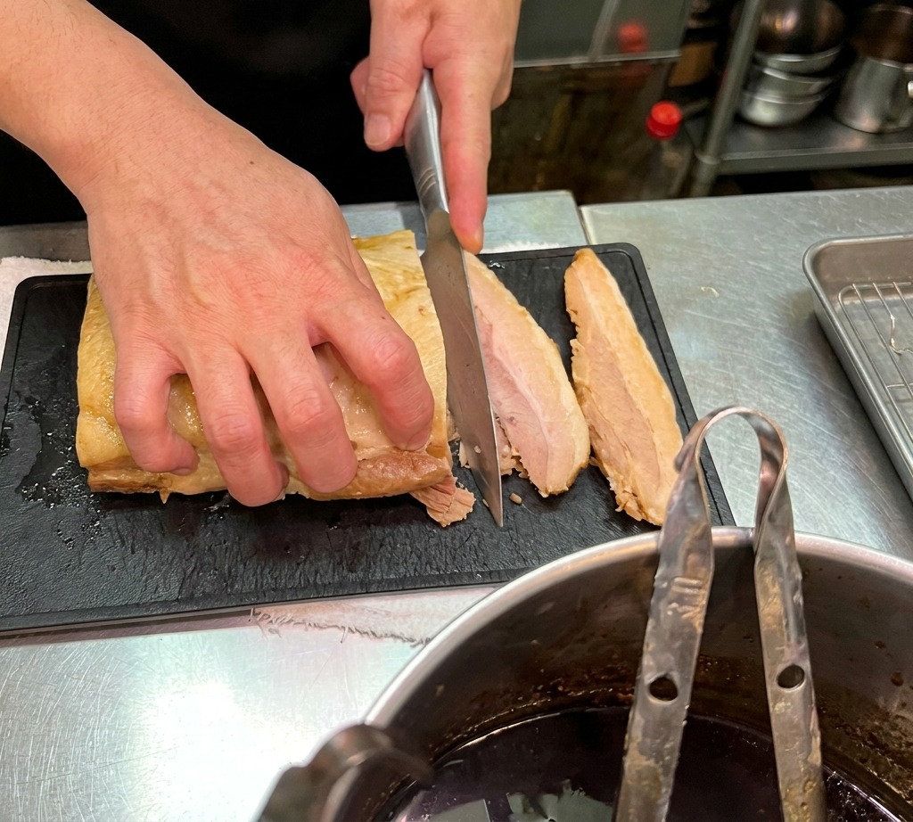 黒いまな板の上で、柔らかく煮込まれた豚バラ肉を包丁でスライスしているクローズアップ。