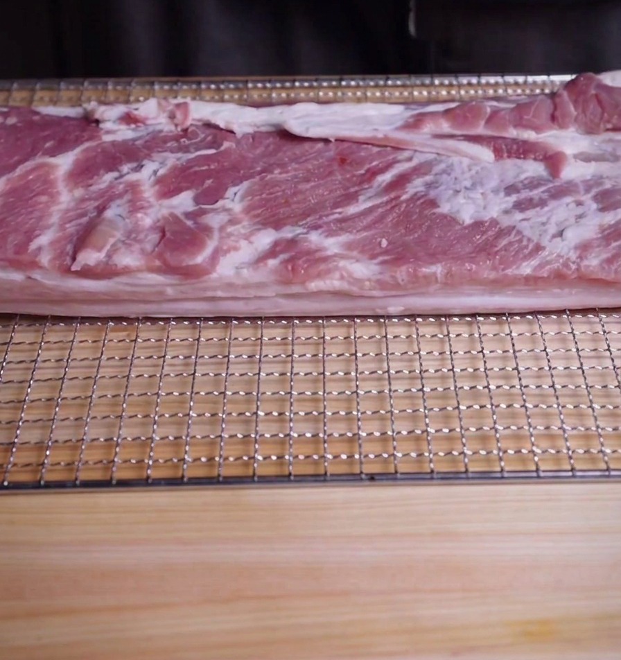 木製のまな板とワイヤーラックの上に置かれた、大きな長方形の豚バラ肉のブロック。