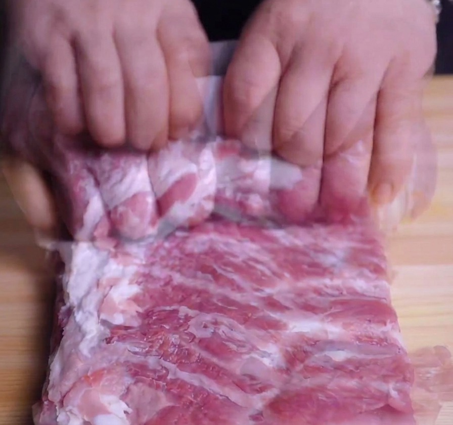木製のまな板の上で、豚バラ肉をきつく円筒状に巻いている手元のアップ。