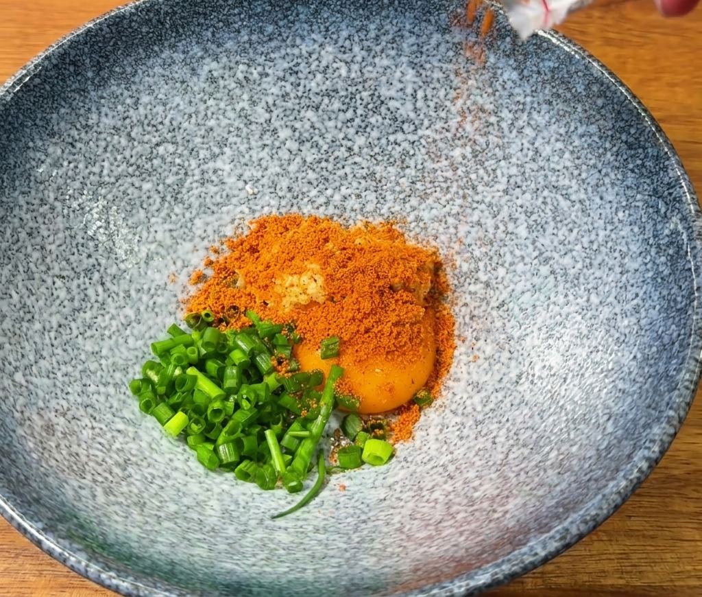 青い器に卵黄、刻みねぎ、赤いラーメンの粉末スープを入れている様子。