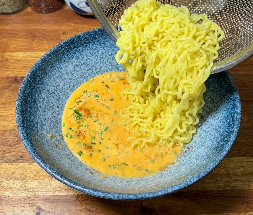 ザルで湯切りした麺を、オレンジ色のスープが入った器に移している様子。