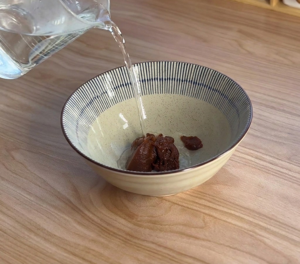 丼の中の味噌ペーストに向かって、計量カップから熱々のお湯を注いでいる様子。