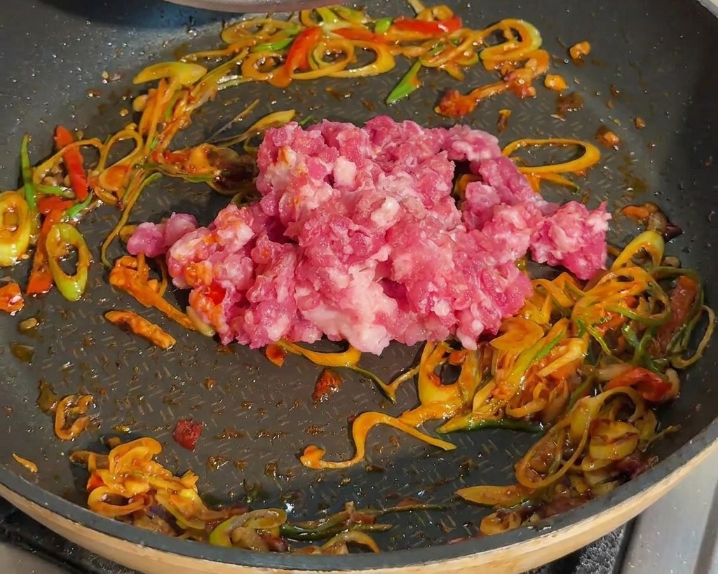 ねぎと豆板醤を炒めたフライパンに、生の豚ひき肉を加える様子。