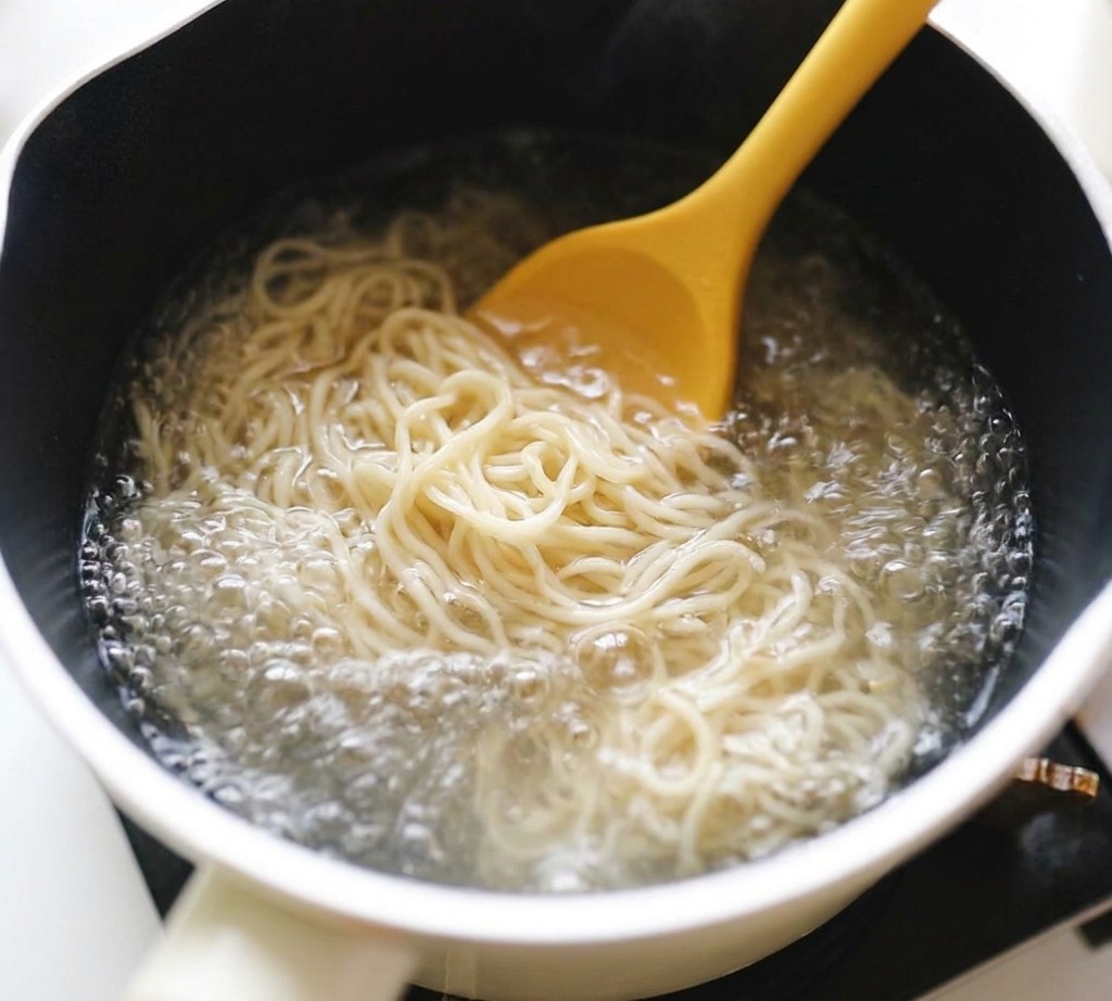 沸騰した鍋の中で、縮れのあるラーメンの麺を箸でほぐしながら茹でている様子。