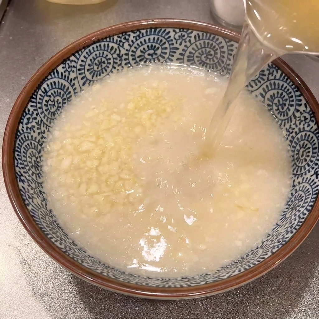 おろしたてのニンニクが入った青と白のラーメン鉢に、鍋から熱いスープを注いでいる様子。