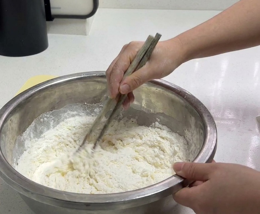 ステンレスボウルの中で、木製のお箸を使って小麦粉と冷水を混ぜ、そぼろ状の生地を作る様子。