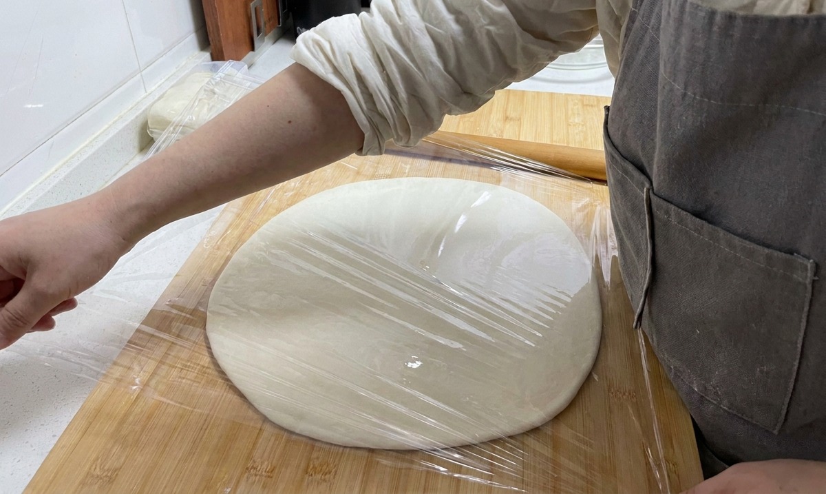 木製ボードの上で、平らに広げられた生地に透明なラップをかけて密閉する手。