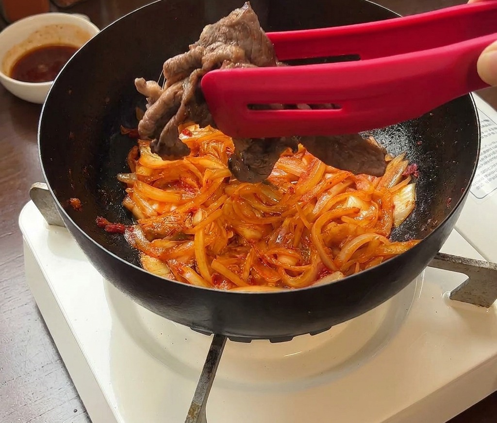 炒めた玉ねぎとキムチの入ったフライパンに、取り出しておいた牛肉を戻し入れる様子。