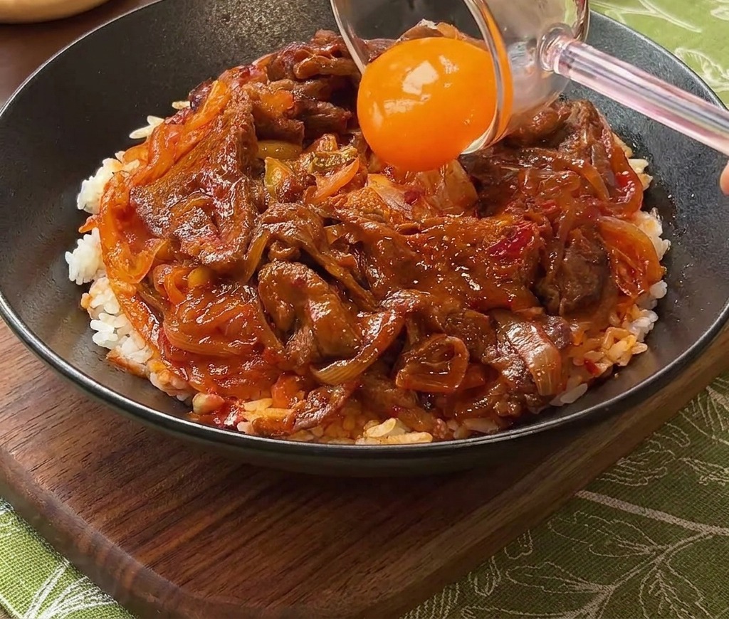 キムチ牛丼の中央に、鮮やかなオレンジ色の卵黄を乗せる様子。