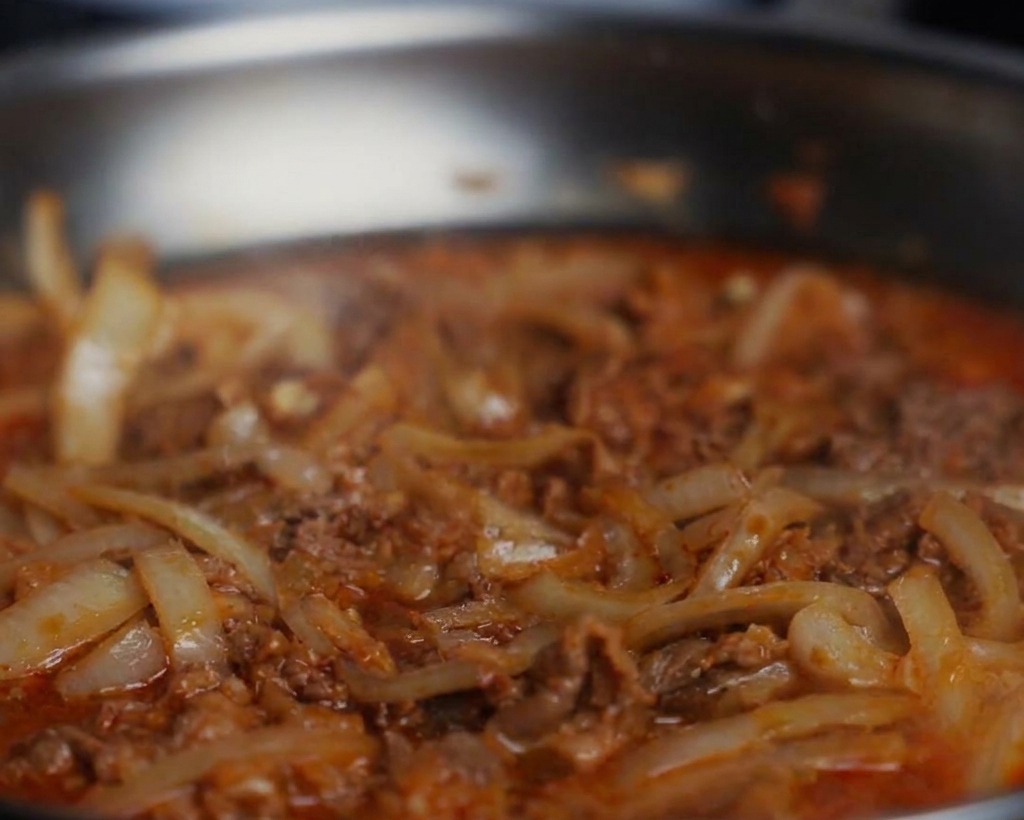 グツグツと煮立っている牛肉と玉ねぎのフライパンに、黒こしょうと醤油を加えているクローズアップ写真。