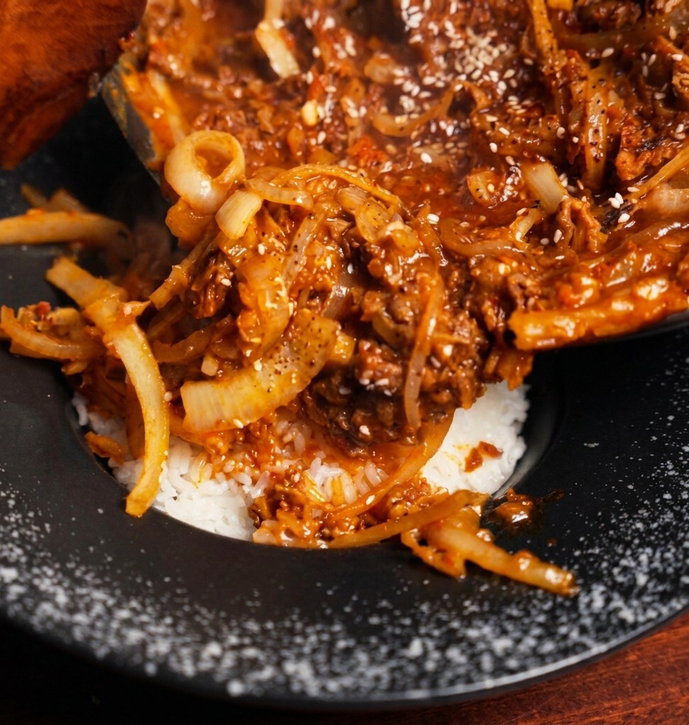 黒い丼に盛られたホカホカの白いご飯の上に、濃厚な牛肉と玉ねぎの炒め物をフライパンから直接のせている様子。