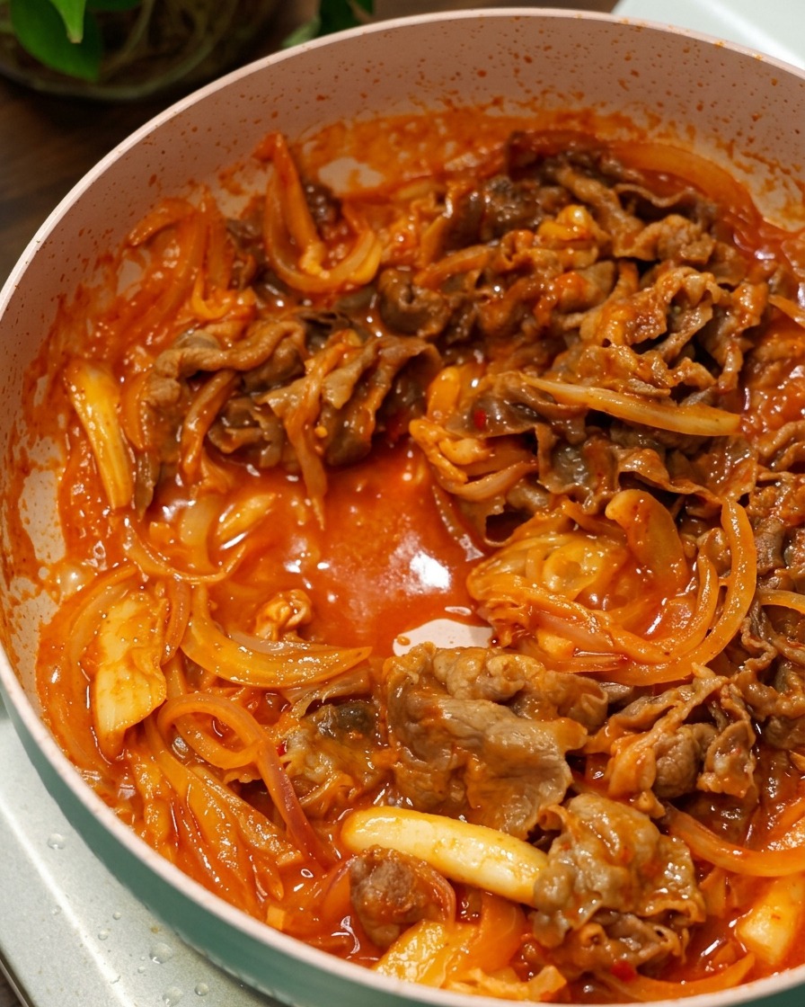 ピンク色のフライパンの中で、牛肉と玉ねぎがグツグツと煮立つ赤い旨辛ソースに絡んでいる様子。