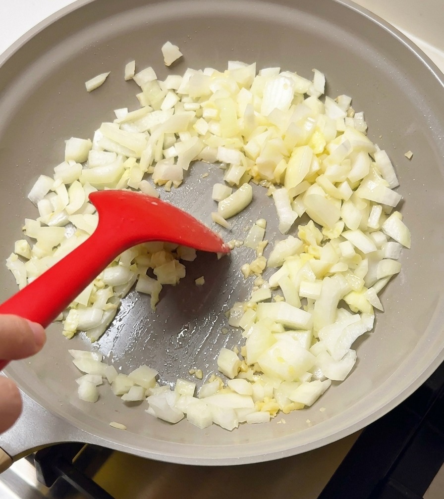 薄灰色のフライパンで、みじん切りにした白玉ねぎとにんにくを赤いシリコンスパチュラで炒めている様子。