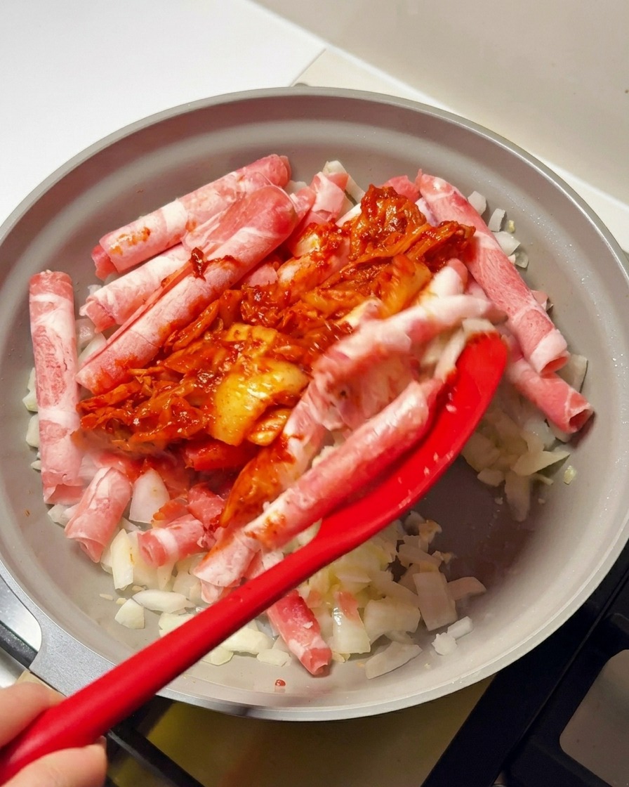 フライパンの牛肉と玉ねぎに、赤いキムチとコチュジャンのソースをスパチュラで加えている様子。