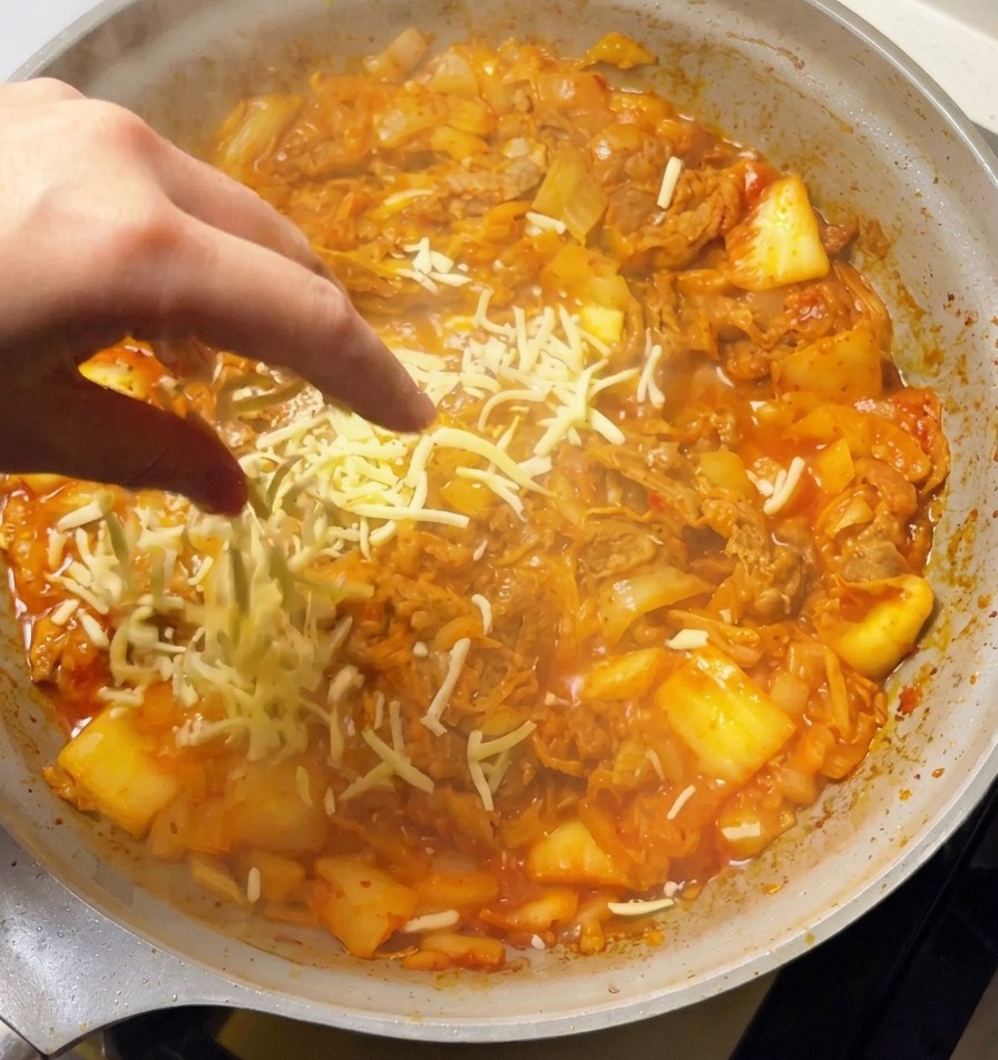 グツグツと煮立つスパイシーなキムチと牛肉の炒め物の上に、ピザ用チーズをたっぷりと振りかけている手。