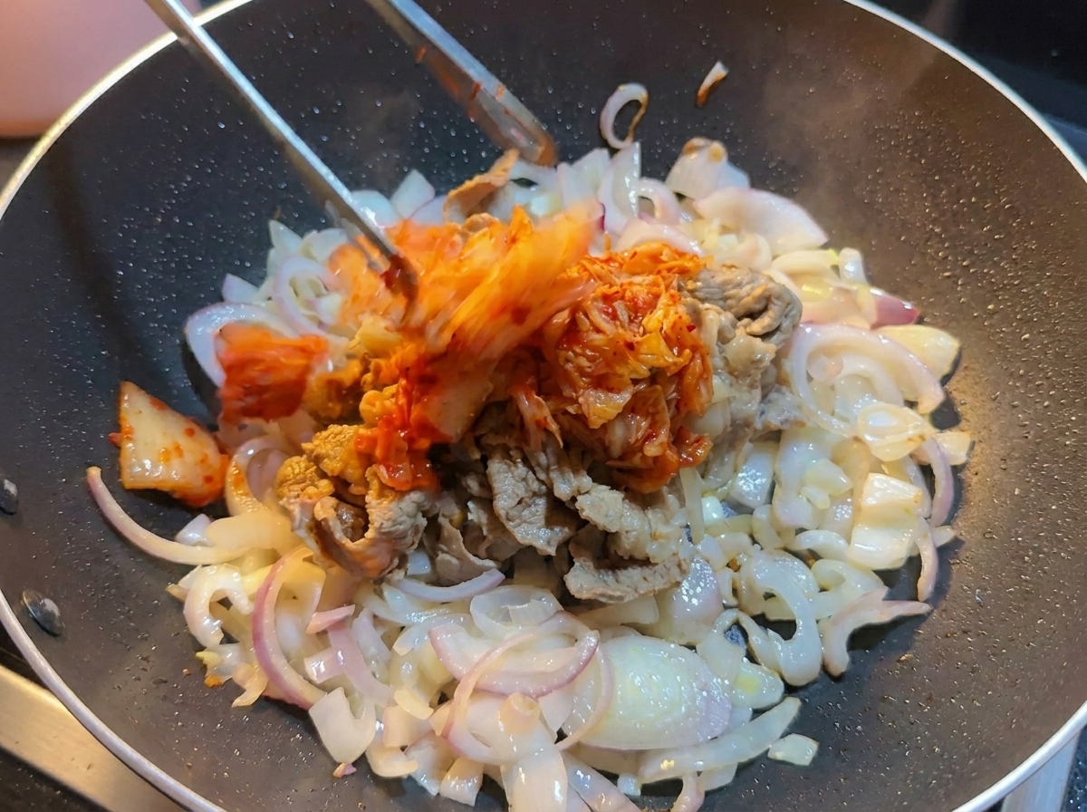 牛肉と玉ねぎが入った鍋に、トングを使って刻んだ白菜キムチを加えている様子。
