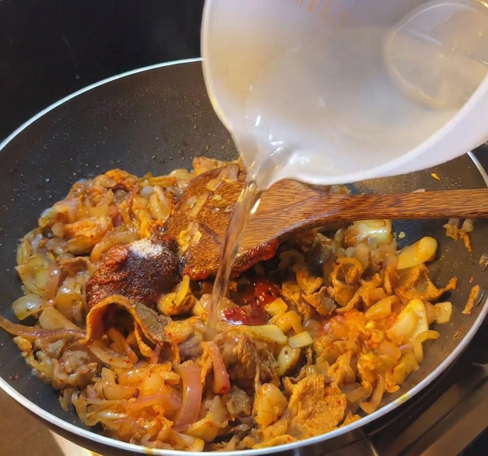 牛肉とキムチの赤いソースが入ったフライパンに、白い器から水を注いでいる様子。