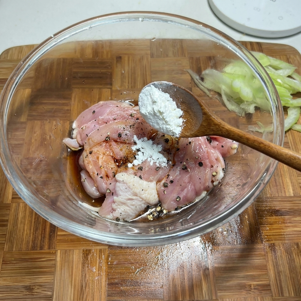 ボウルに入った味付け済みの鶏肉に、木のスプーンで片栗粉を加えているクローズアップ写真。