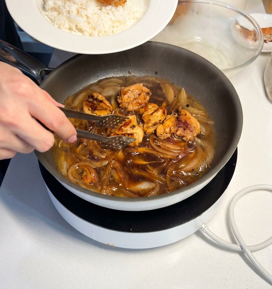 炊きたての白いご飯の上に、トングを使って照りよく煮上がった鶏肉を盛り付けているシーン。